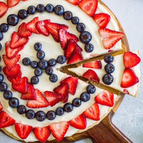 Fourth of July fruit pizza on a round wooden cutting board with strawberries surrounding it.