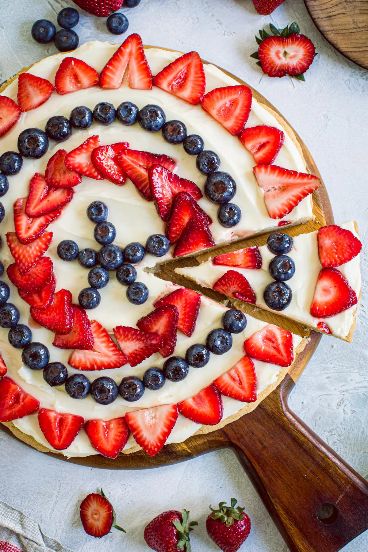 Fourth of July fruit pizza on a round wooden cutting board with strawberries surrounding it.