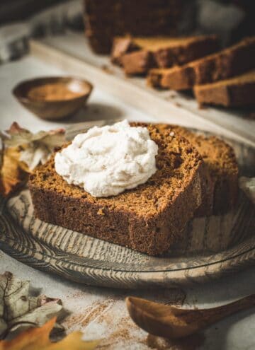 Moist pumpkin bread slice topped with whipped cream on a wooden plate.
