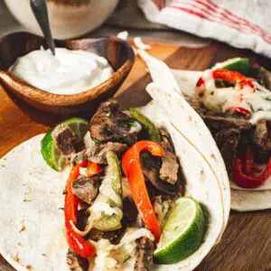 Cheesesteak fajitas on a round board with sour cream and cheese in bowls behind.