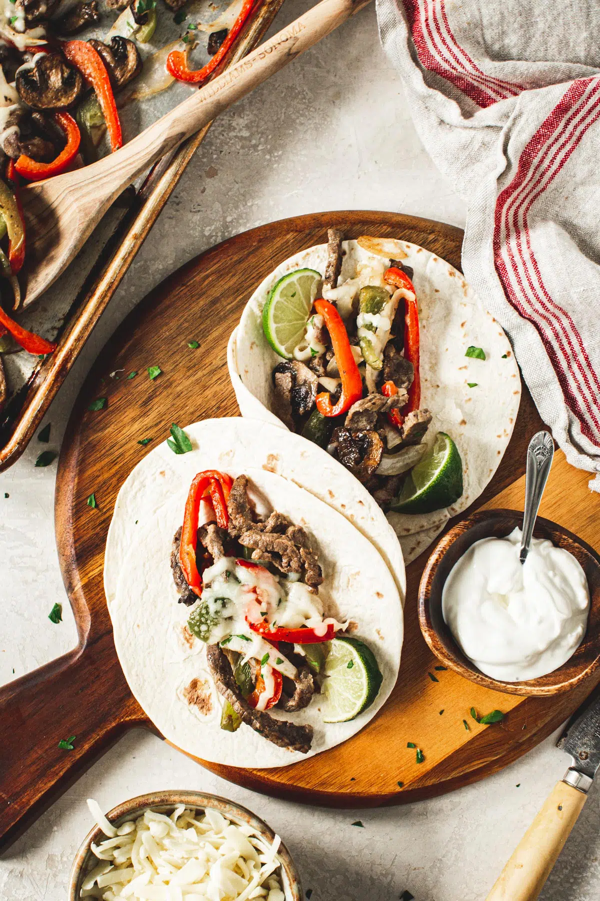 Homemade cheesesteak fajitas on a round wooden board with sour cream in a bowl beside it.