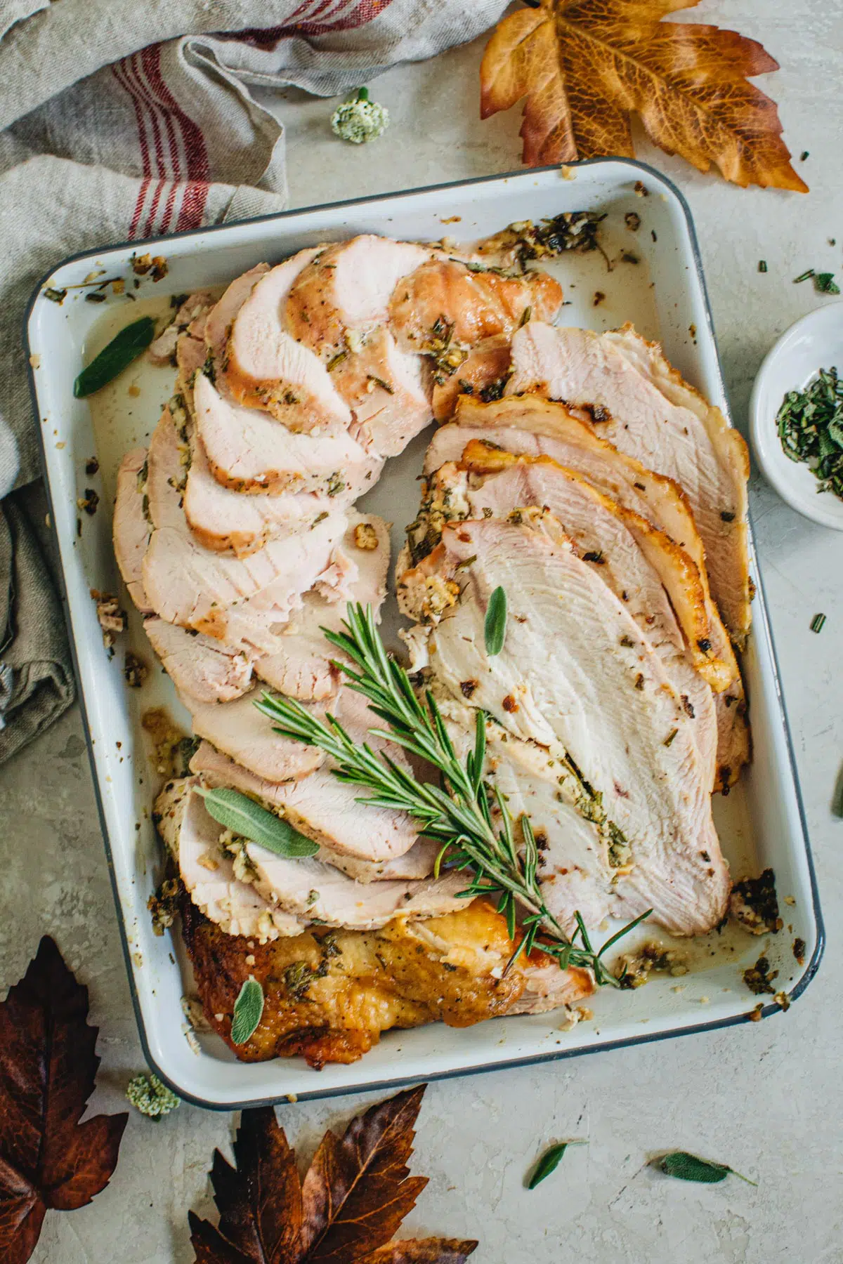 Oven-roasted turkey breast sliced on a rimmed baking sheet and topped with fresh rosemary and sage.