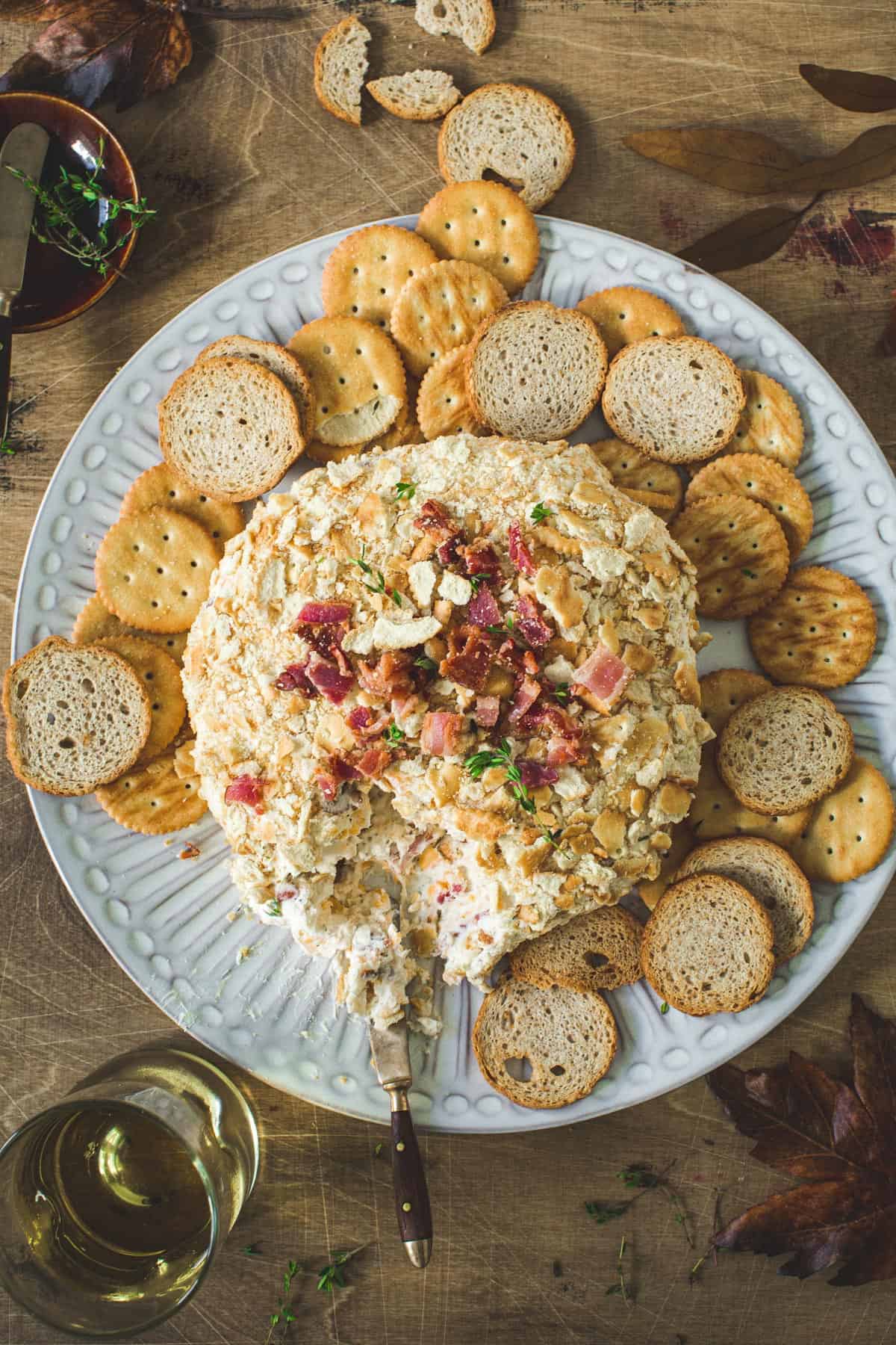 Bacon cheddar cheese ball on a white round plate with crackers and melba toast surrounding it.