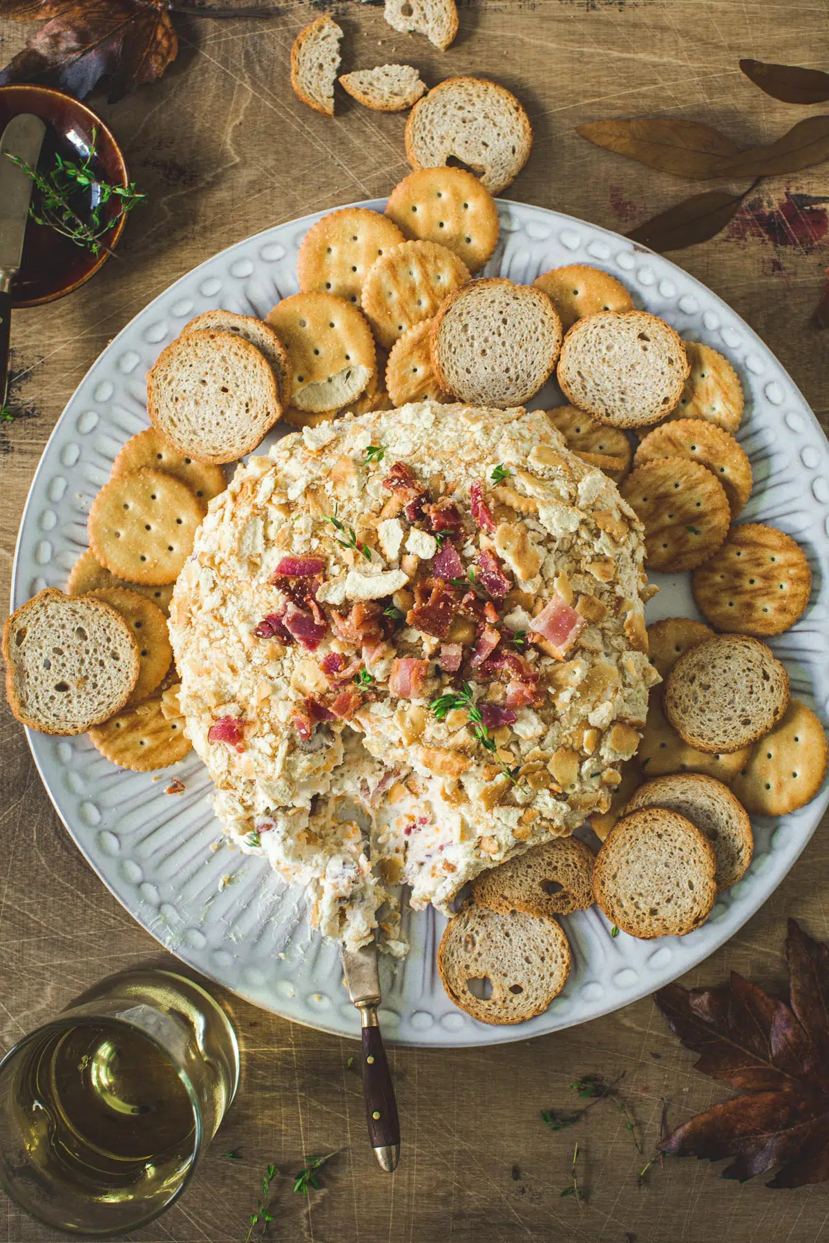 Bacon cheddar cheese ball on a white round plate with crackers and melba toast surrounding it.
