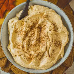 Pumpkin cheesecake dip with cinnamon sprinkled on top in a bowl with a wooden spoon.