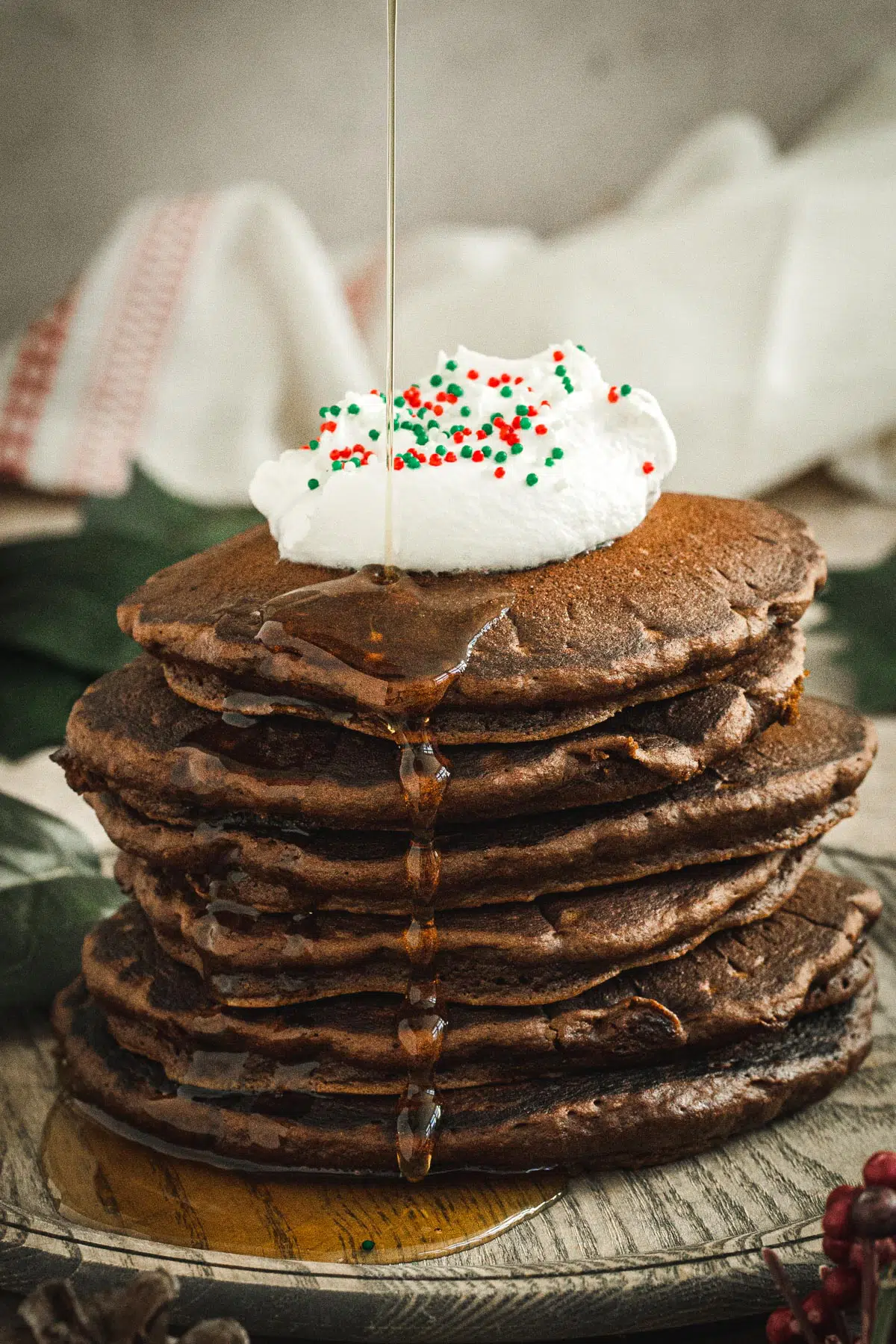 Stack of gingerbread pancakes topped with whipped cream and sprinkles with syrup dripping down the side.