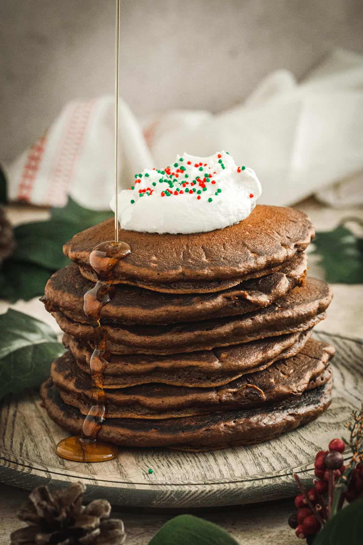 Stack of gingerbread pancakes topped with whipped cream and sprinkles and a drizzle of maple syrup dripping down the side.