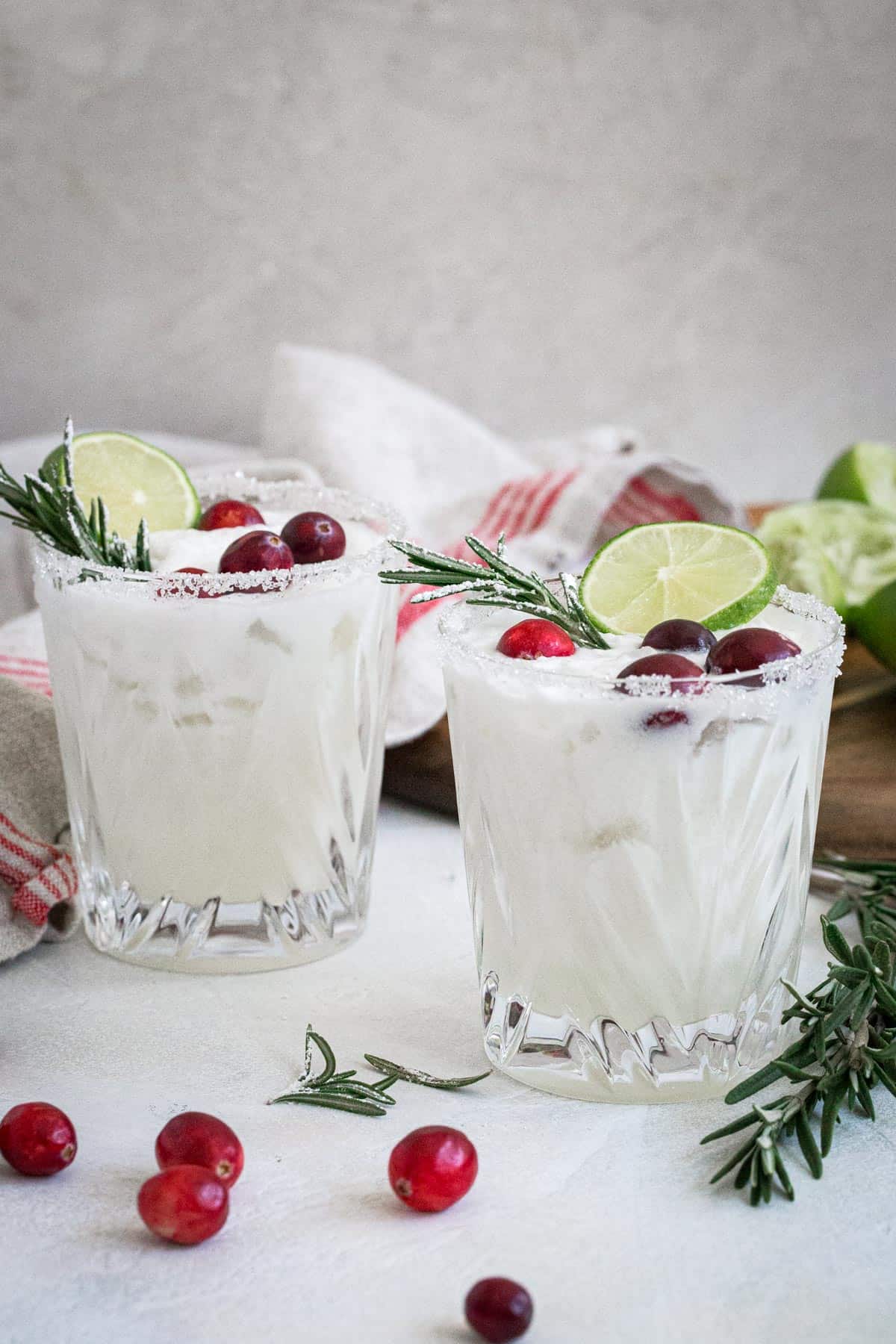 Two white Christmas margaritas topped with lime slice, fresh cranberries, and sugared rosemary.