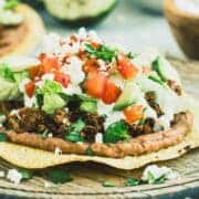 Beef tostada topped with cheese, diced tomatoes, and avocado on a wooden plate.