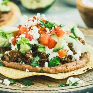 Beef tostada topped with cheese, diced tomatoes, and avocado on a wooden plate.