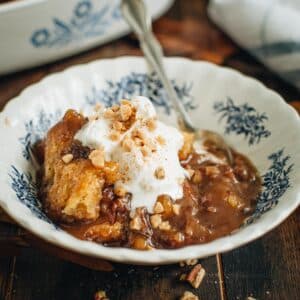 Pecan pie cobbler topped with whipped cream and toffee bits in a blue and white bowl.