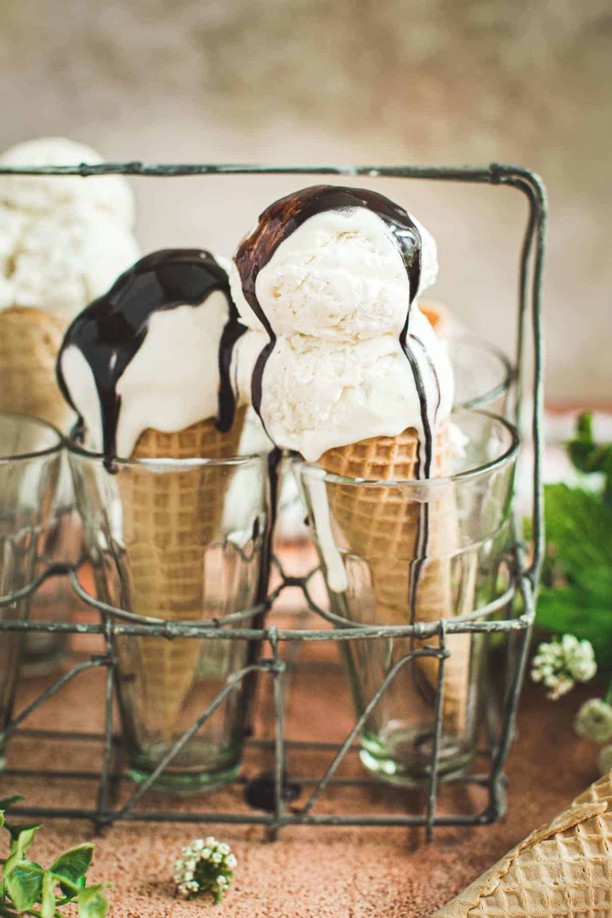 Vanilla bean ice cream in a cone topped with chocolate syrup. Cones are in a glass.
