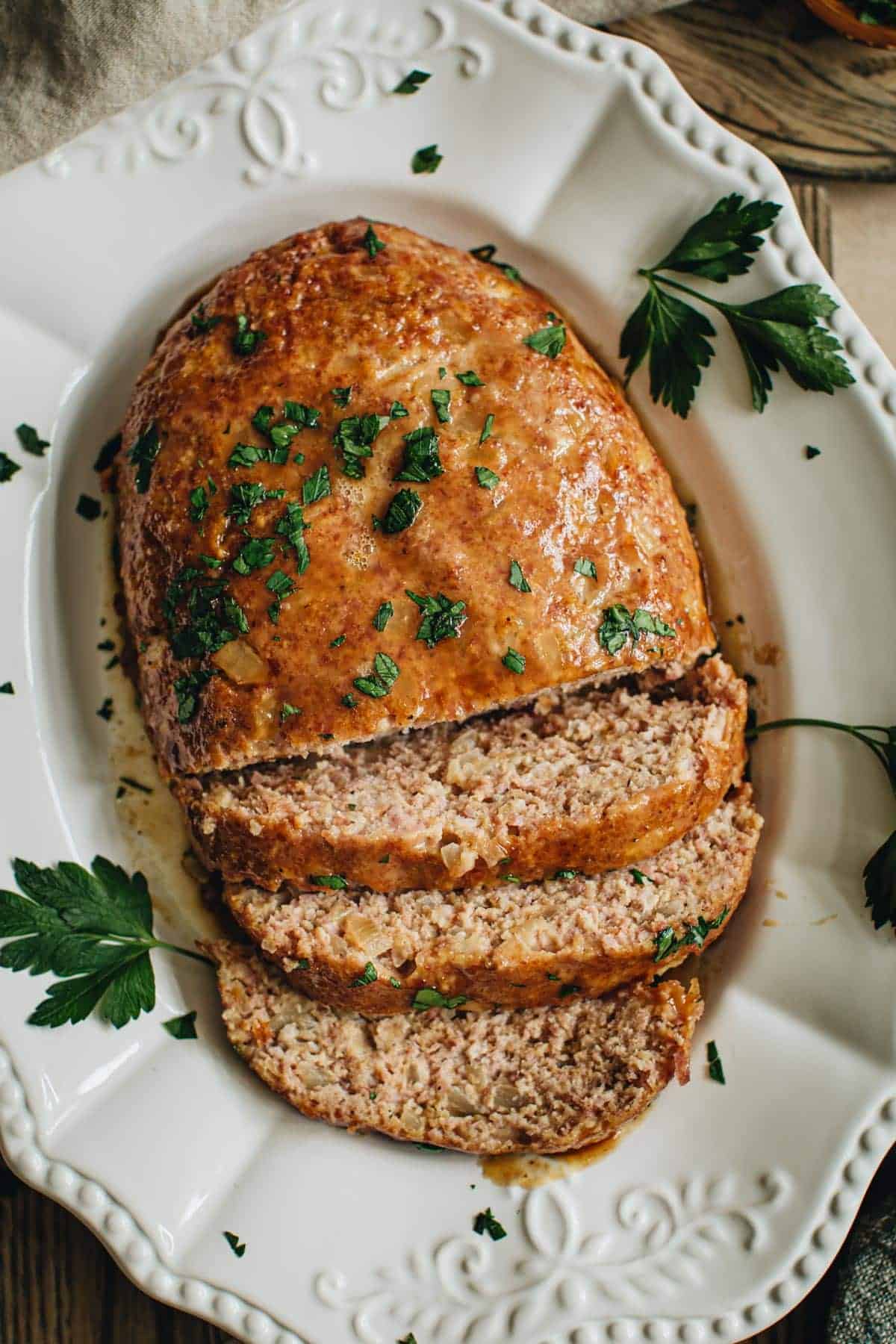 Ham loaf on a white platter with half of it sliced and sprinkled with fresh parsley.