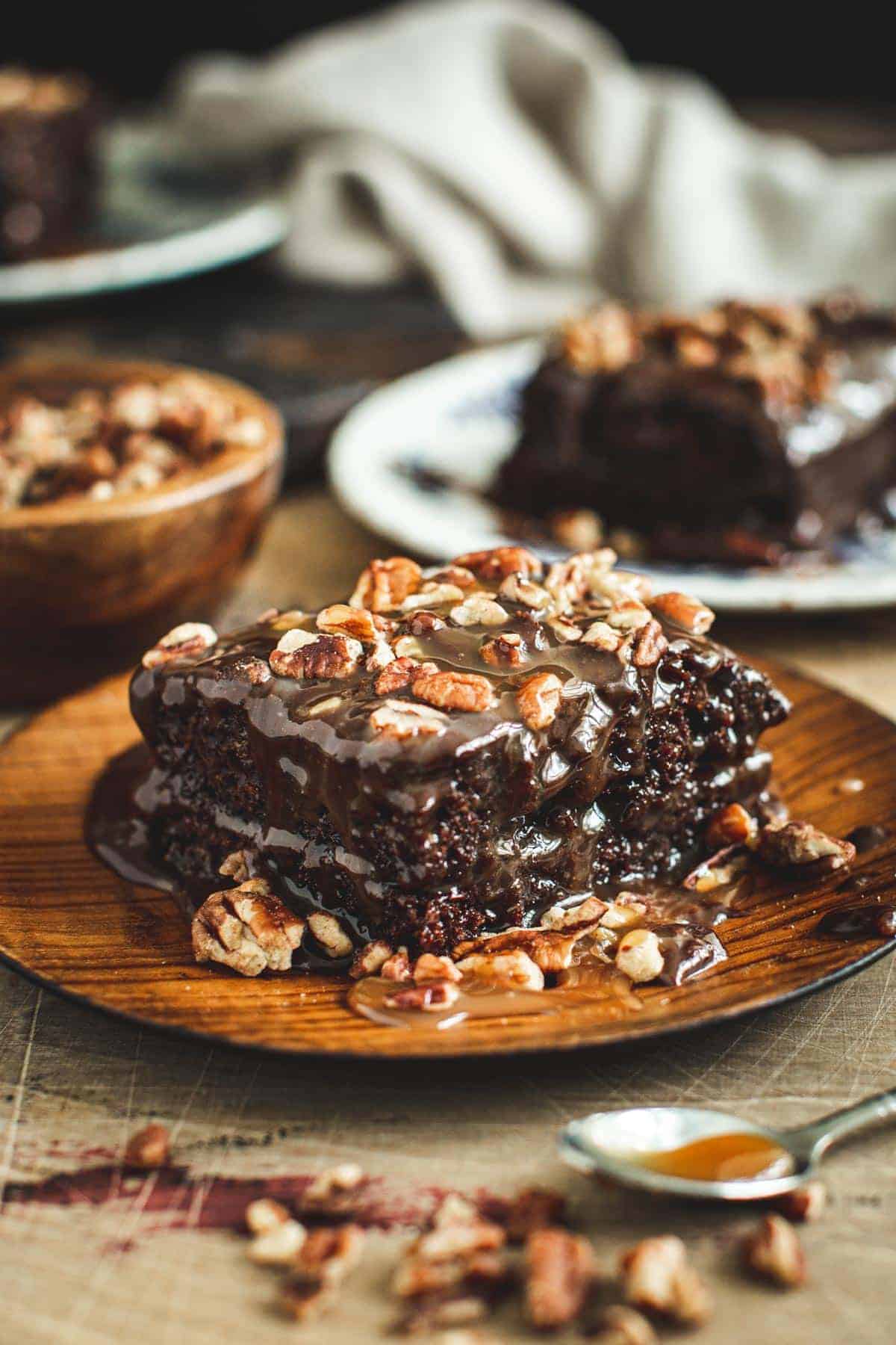 Slice of turtle cake covered with chopped pecans on a wooden plate.