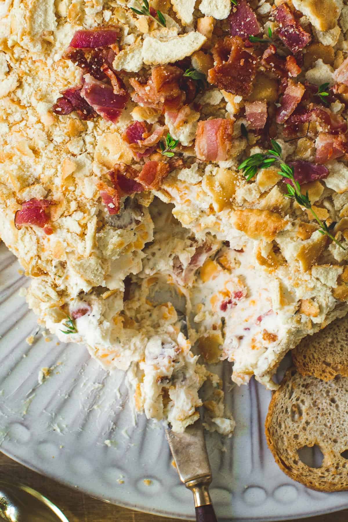 Bacon ranch cheese ball with a portion scooped out on a serving knife with crackers next to it.