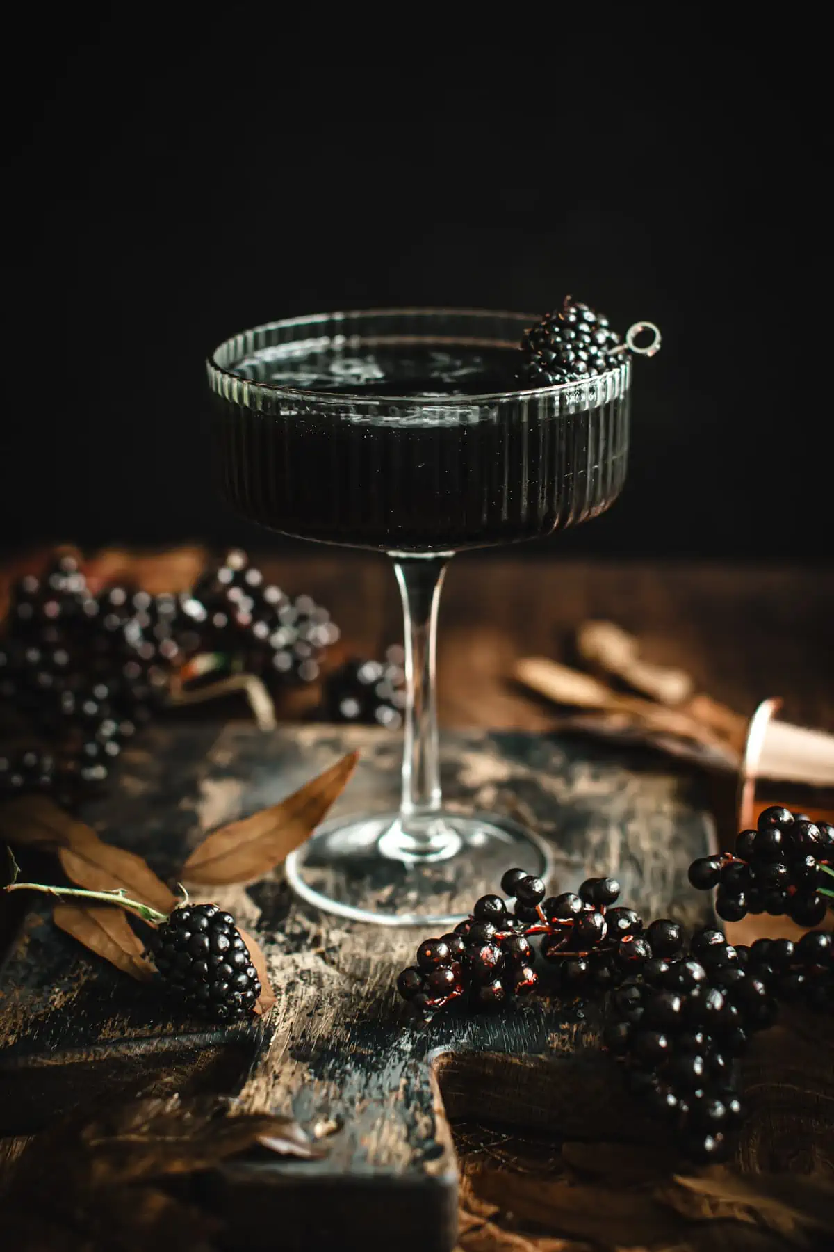 A black martini in a ribbed martini glass with a blackberry garnish on a cocktail pick.