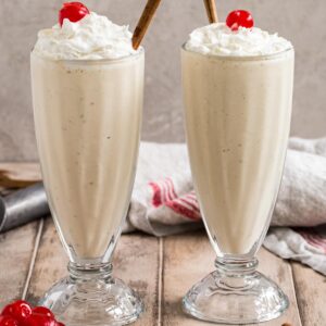 Eggnog milkshakes topped with whipped cream, cherries, and cinnamon sticks in a milkshake glass.