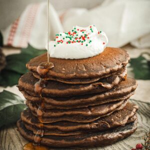 Gingrebread pancakes stacked with whipped cream on top and syrup dripping down the side.