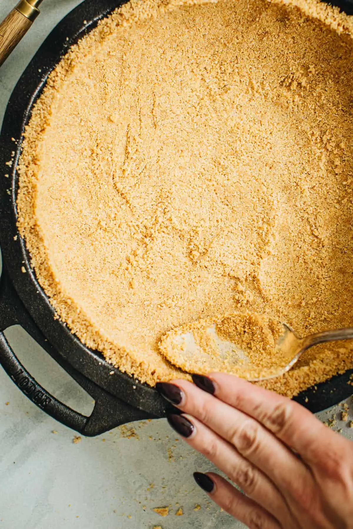 Hand and spoon pressing the edges of a graham cracker crust to shape it.