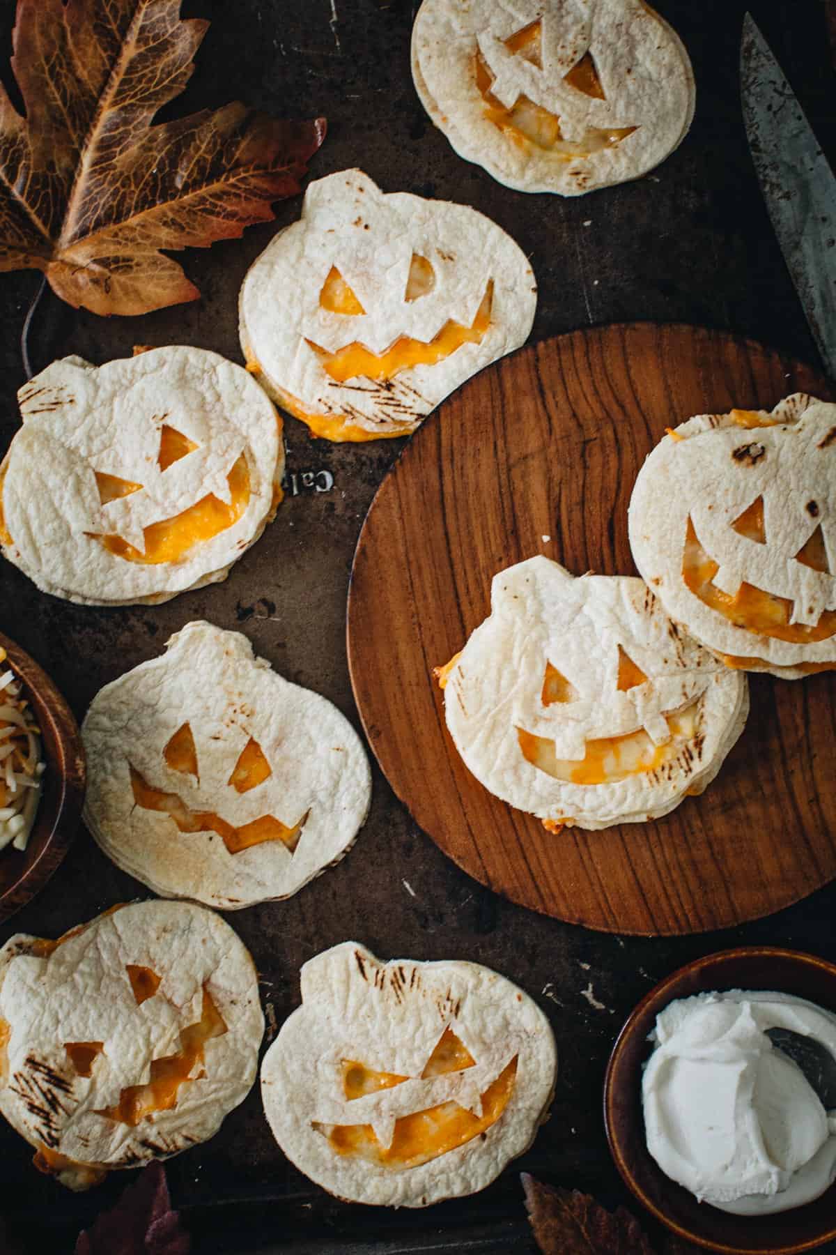 Jack-O-Lantern quesadillas spread on a woode plate and baking sheet with toppings surrounding.