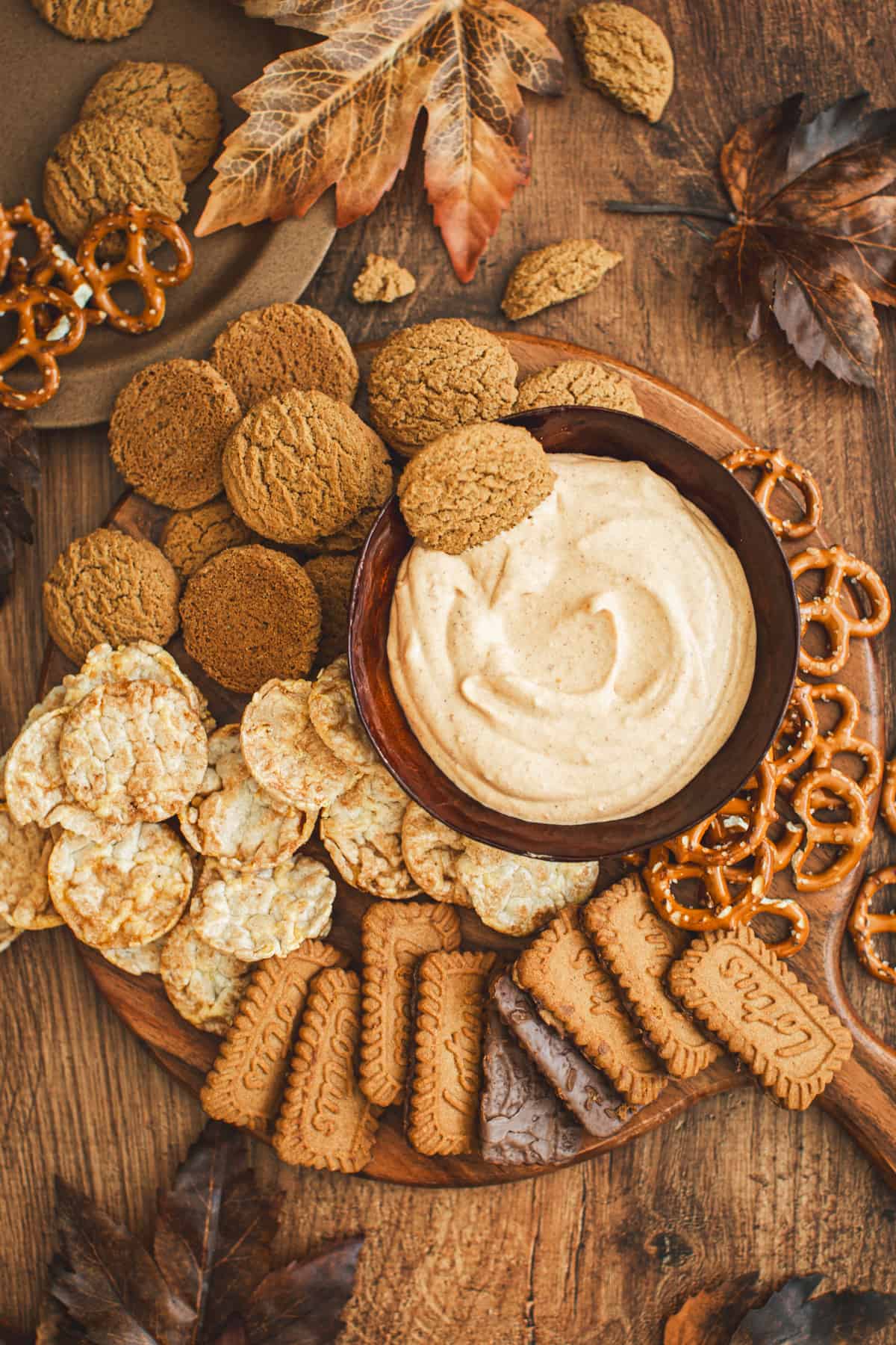 Pumpkin dip in bowl sitting on a board with cookies, pretzels, and rice cakes around it.