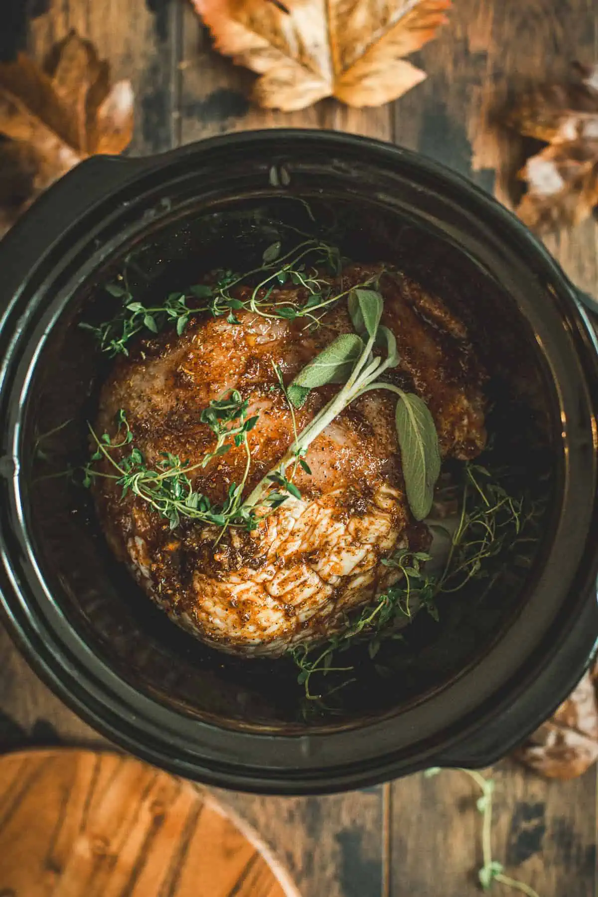 Slow cooker with turkey breast covered in rub and fresh thyme and sage on top.