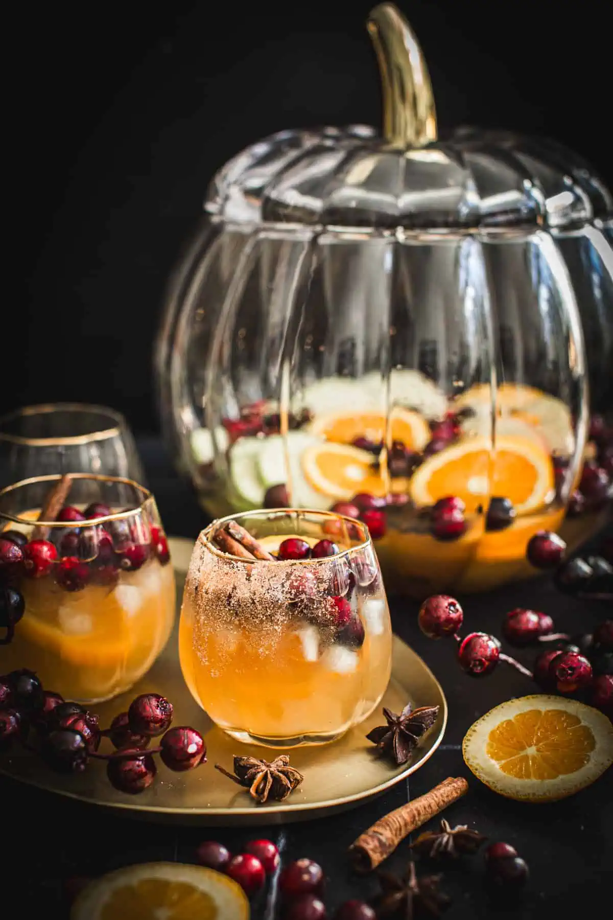 Thanksgiving sangria in a pumpkin-shaped glass with a pumpkin-shaped punch bowl filled with sangria behind it.