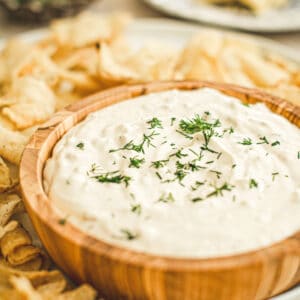 Clam dip topped with fresh dill in a wooden bowl with chips around it.