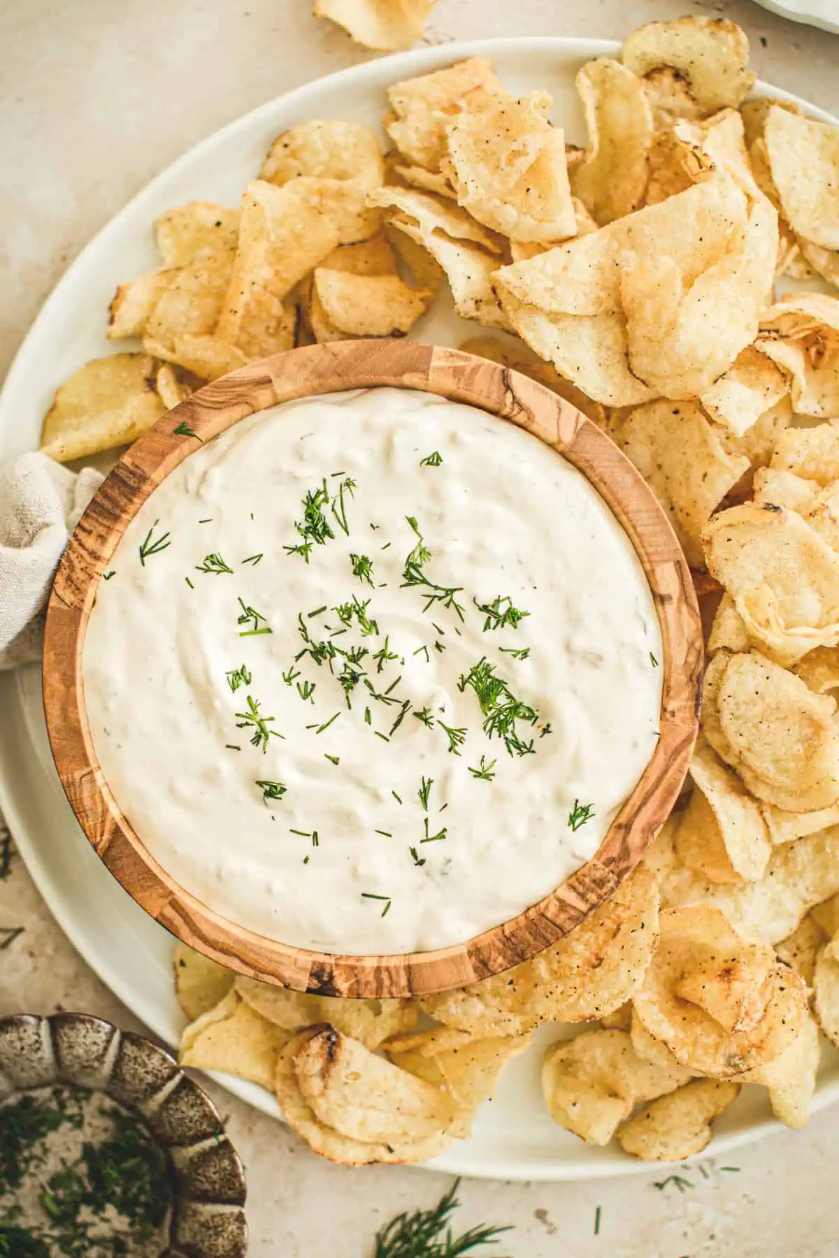 Clam dip sprinkled with fresh chopped dill in a wooden bowl on a round plate with chips around it.