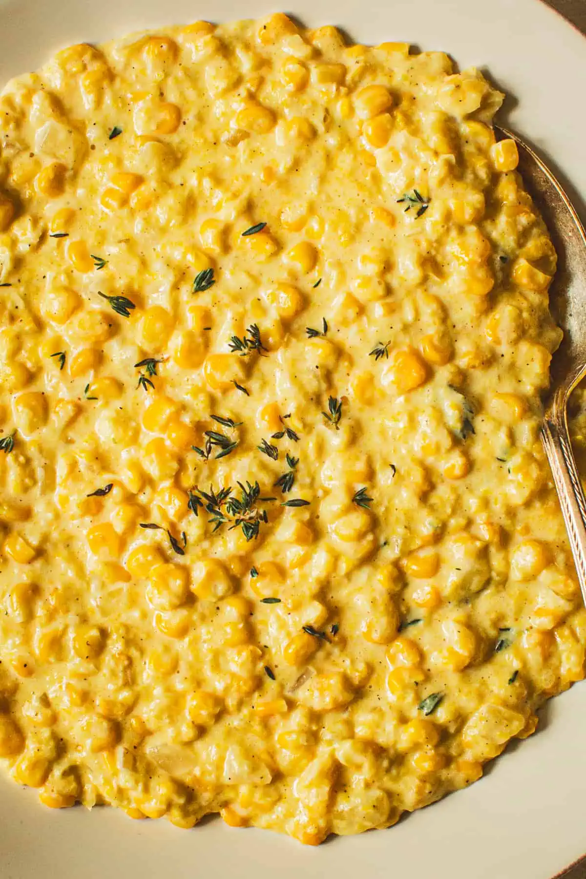 Creamed corn topped with fresh thyme in a white bowl with a silver serving spoon.