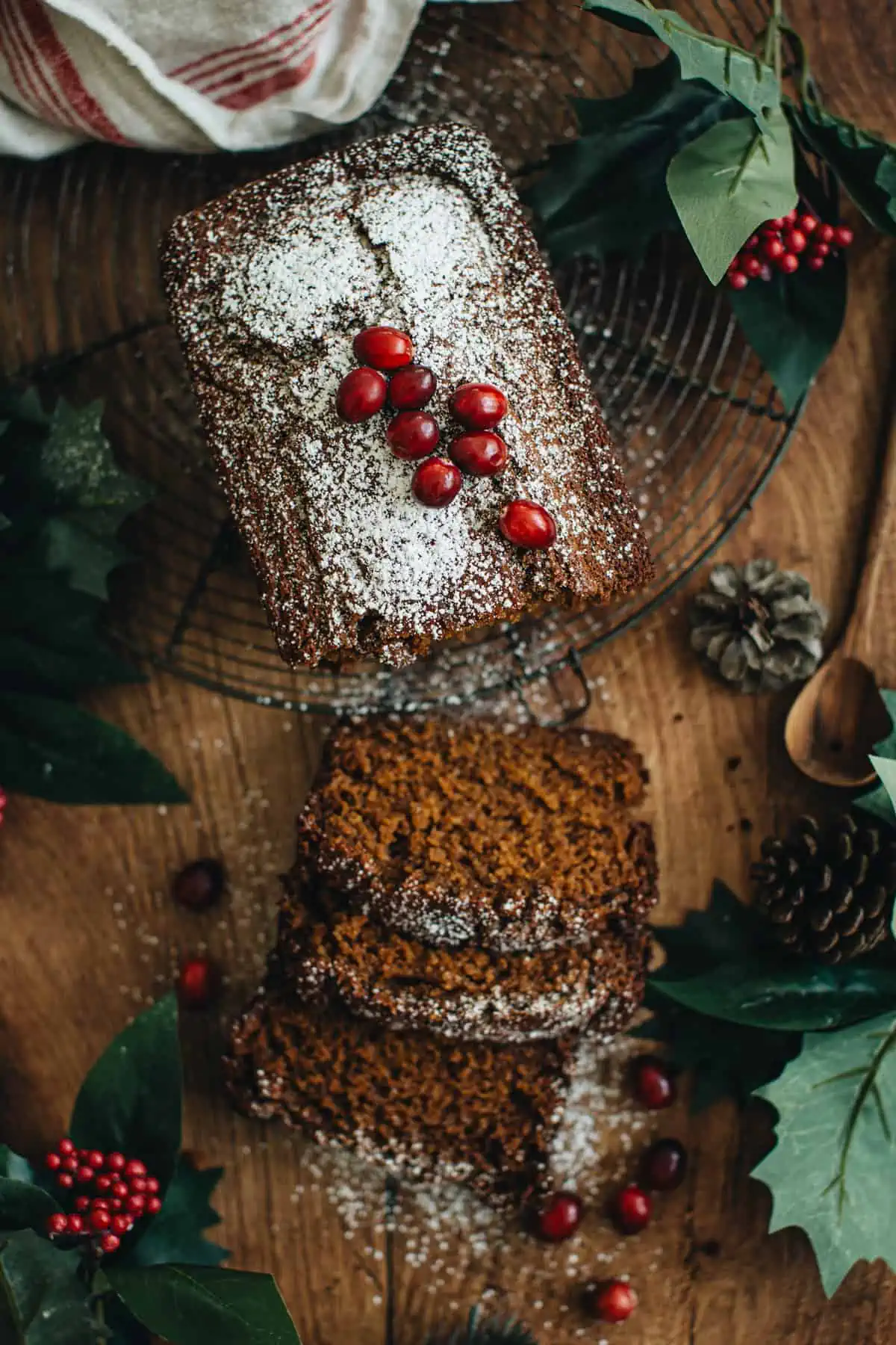 Gingerbread loaf dusted with powdered sugar and topped with fresh cranberries sitting on a round wire rack. First three pieces of the loaf are sliced and sitting next to it.