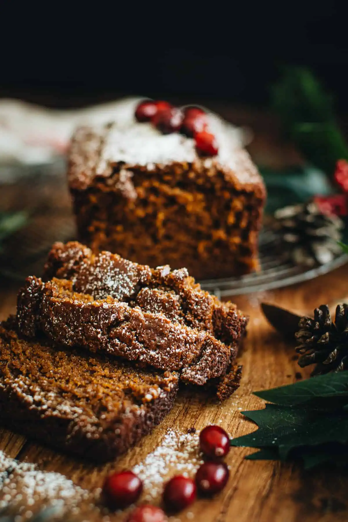 Gingerbread loaf dusted with powdered sugar on a round wire rack with three slices cut from the front. Christmas greenery around the slices and loaf as well as fresh cranberries.