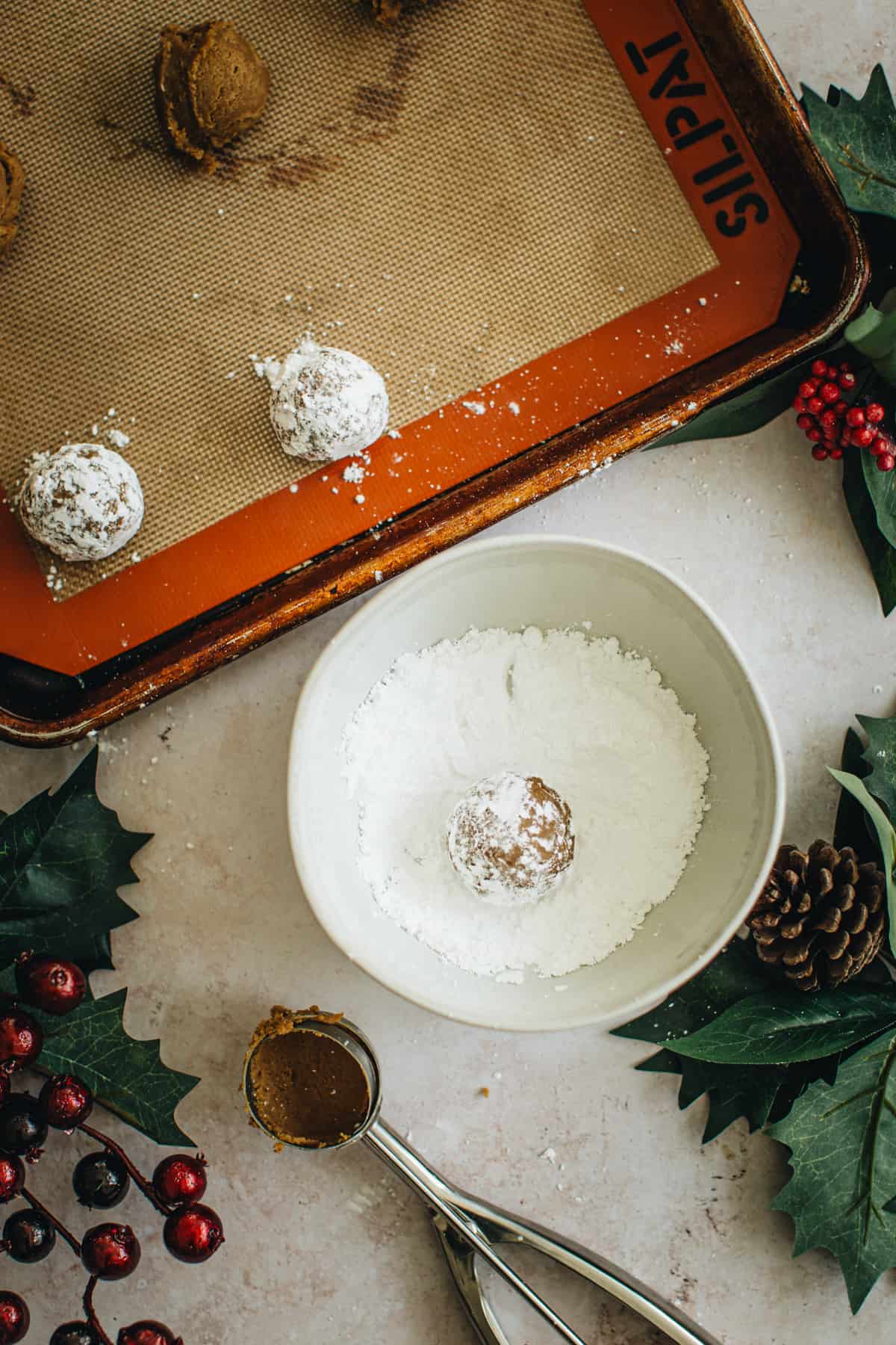 Molasses crinkle cookie dough balls in a baking sheet and one rolled in a bowl of powdered sugar.