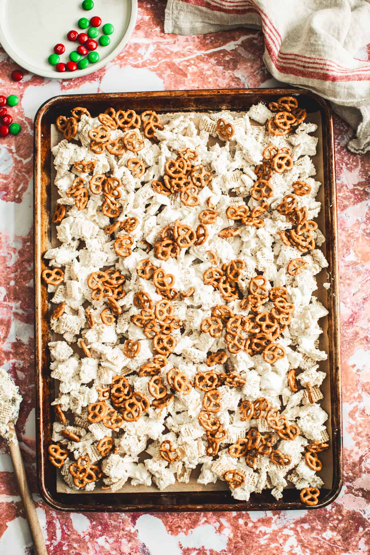 Pretzels sprinkled on top of white chocolate coated cereal on a parchment-lined baking sheet for making Christmas Chex mix.