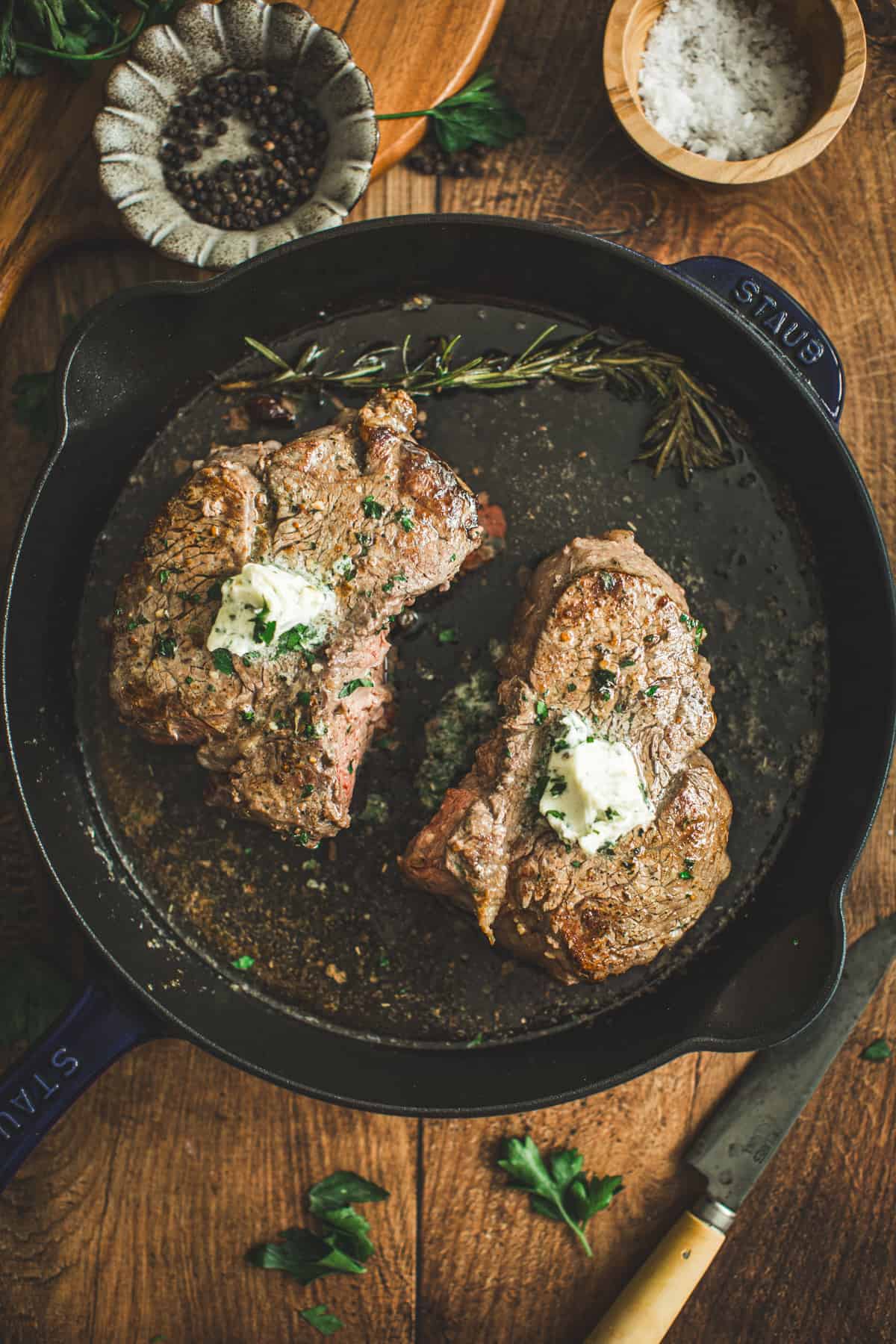 Two pan-seared filet mignon filets in a cast iron skillet topped with herb butter and fresh parsley.