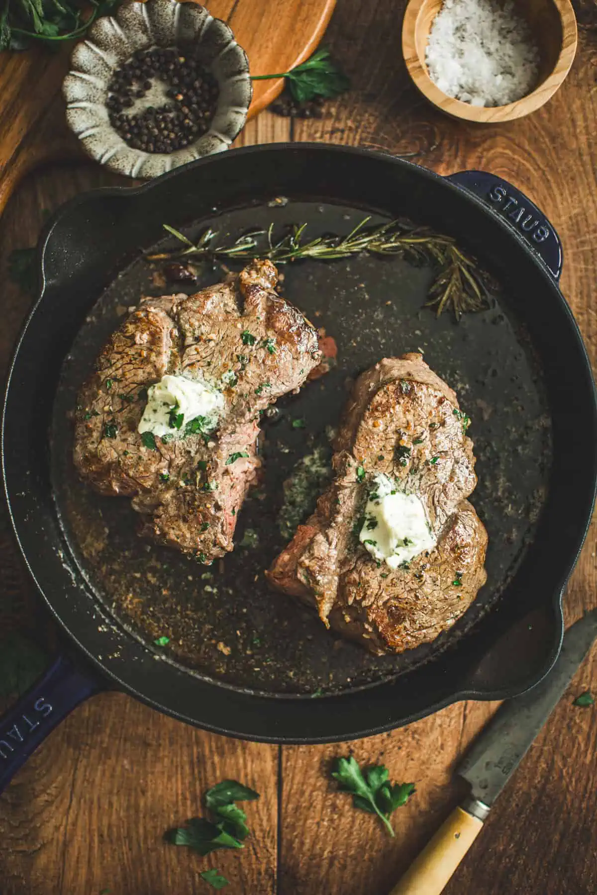 Two pan-seared filet mignon filets in a cast iron skillet topped with herb butter and fresh parsley.