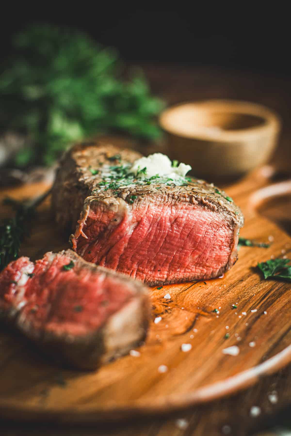 Pan-seared filet mignon with a slice cut off and topped with herb butter. Sitting on a round wooden cutting board.