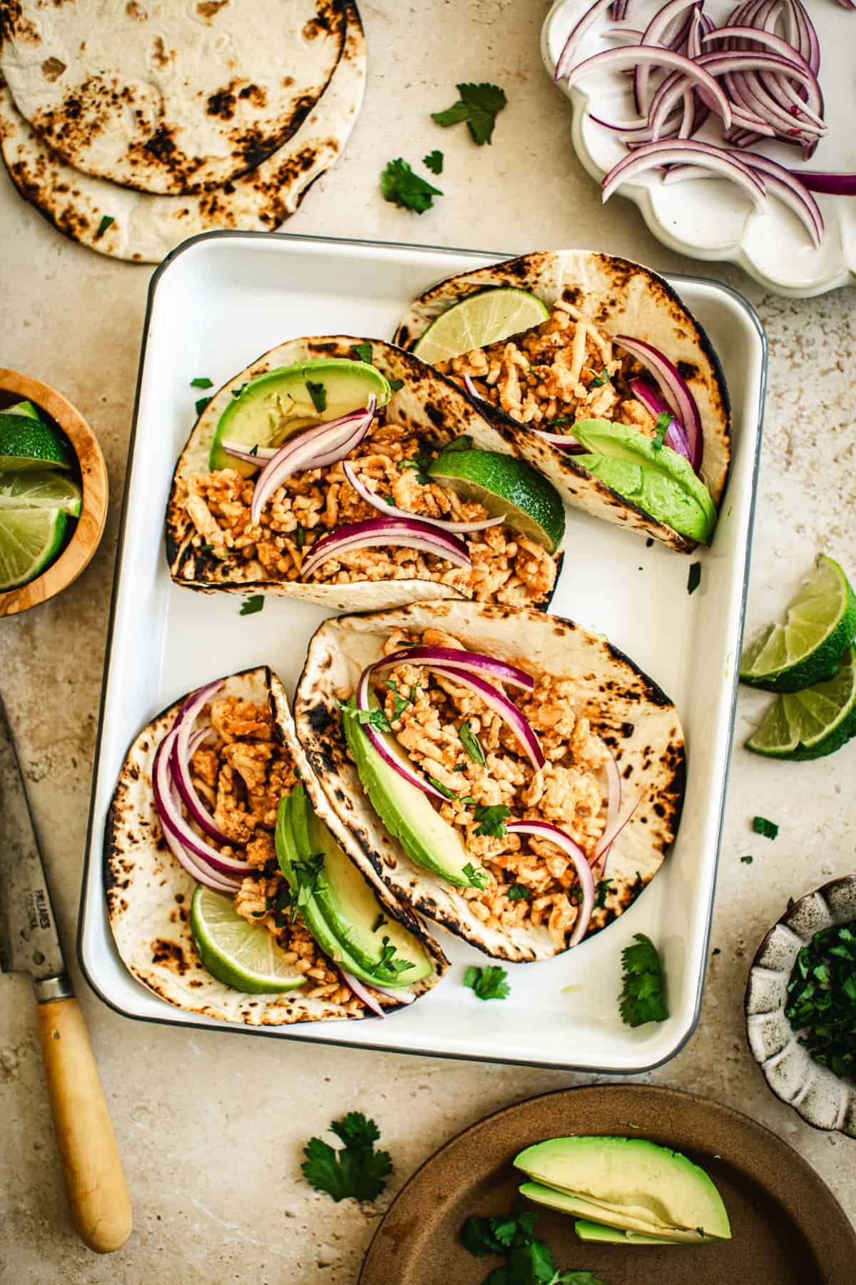 Ground chicken tacos on a tray topped with avocado, red onions, and lime wedges.